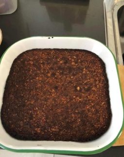 A square white enamel baking dish with a green rim, holding a dark golden-brown baked coconut and almond slab, on a dark benchtop.