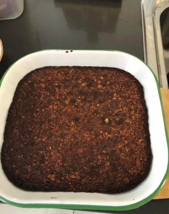 A square white enamel baking dish with a green rim, holding a dark golden-brown baked coconut and almond slab, on a dark benchtop.