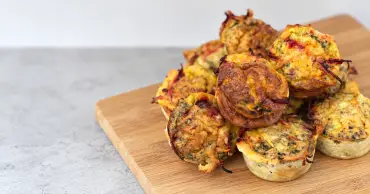 Homemade beetroot egg muffins with golden cheese tops and visible vegetable shreds, stacked on a wooden chopping board.