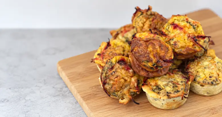 Homemade beetroot egg muffins with golden cheese tops and visible vegetable shreds, stacked on a wooden chopping board.