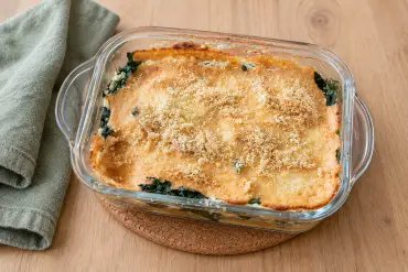 A golden ricotta bake with spinach showing at the edges, in a glass dish on a cork mat beside a green tea towel.