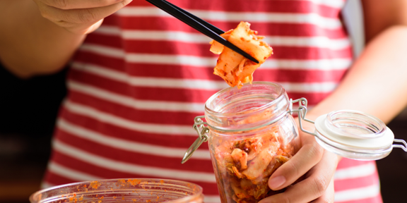 Lifting a forkful of kimchi out of a glass jar with chopsticks.