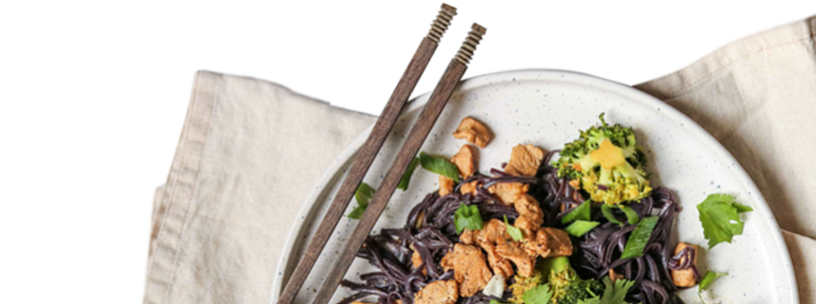Bowl of black noodles topped with browned protein pieces, broccoli and fresh herbs, with wooden chopsticks.