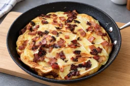 A golden bacon and brie frittata in a black frying pan on a wooden board, topped with crispy chopped bacon and thin slices of melted Brie.