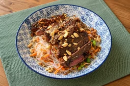 Sliced seared steak on konjac noodles with grated carrot, red onion, edamame and chopped peanuts, in a blue patterned bowl on a green placemat.
