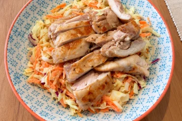 A blue and white patterned bowl with an orange rim, holding creamy slaw of cabbage and carrot topped with sliced, pan-cooked chicken thigh.