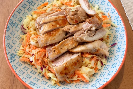 A blue and white patterned bowl with an orange rim, holding creamy slaw of cabbage and carrot topped with sliced, pan-cooked chicken thigh.