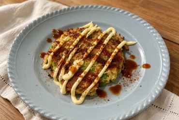 A golden-brown savoury pancake on a pale blue-grey plate, striped with creamy mayonnaise and dark okonomi-style sauce, with flecks of green edamame showing through and a sprinkle of spice around the plate.