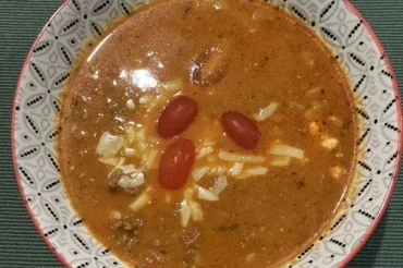A patterned bowl of cheeseburger soup seen from above, with beef mince, grated cheese and three cherry tomatoes on top, on a green placemat.