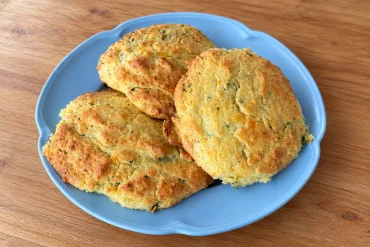 Three golden cheesy keto baps flecked with parsley, stacked on a pale blue plate on a wooden table.