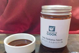 A white label jar of dark brown tonkatsu sauce with a silver lid, beside a small white dish of the sauce on a wooden board, with a red cushion behind.
