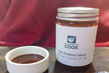 A white label jar of dark brown tonkatsu sauce with a silver lid, beside a small white dish of the sauce on a wooden board, with a red cushion behind.