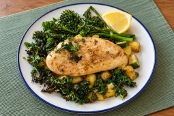 A roasted chicken breast on crispy kale, broccolini, zucchini and potato cubes, with a lemon wedge, on a white plate with a blue rim on a green placemat.