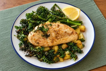 A roasted chicken breast on crispy kale, broccolini, zucchini and potato cubes, with a lemon wedge, on a white plate with a blue rim on a green placemat.