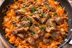 A black frying pan filled with shredded orange vegetables and browned pieces of meat, scattered with chopped parsley and diced onion, on a wooden board beside a striped tea towel.