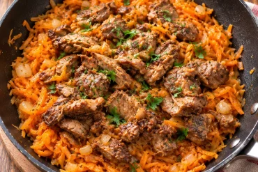 A black frying pan filled with shredded orange vegetables and browned pieces of meat, scattered with chopped parsley and diced onion, on a wooden board beside a striped tea towel.