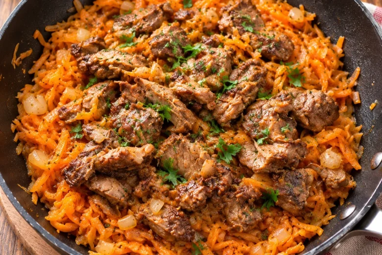 A black frying pan filled with shredded orange vegetables and browned pieces of meat, scattered with chopped parsley and diced onion, on a wooden board beside a striped tea towel.
