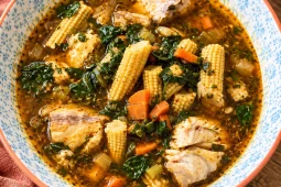 A blue-patterned bowl of spiced fish soup with chunks of white fish, baby corn spears, diced carrot and dark green leaves, on a wooden table.