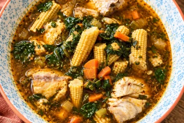 A blue-patterned bowl of spiced fish soup with chunks of white fish, baby corn spears, diced carrot and dark green leaves, on a wooden table.