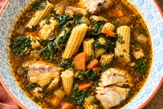 A blue-patterned bowl of spiced fish soup with chunks of white fish, baby corn spears, diced carrot and dark green leaves, on a wooden table.