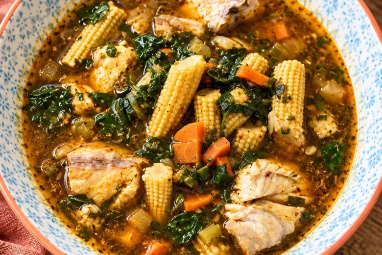 A blue-patterned bowl of spiced fish soup with chunks of white fish, baby corn spears, diced carrot and dark green leaves, on a wooden table.