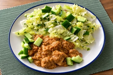 A white plate with a blue rim holding chicken pieces in a thick peanut satay sauce, with shredded pale green leaves and diced cucumber, on a green woven placemat.