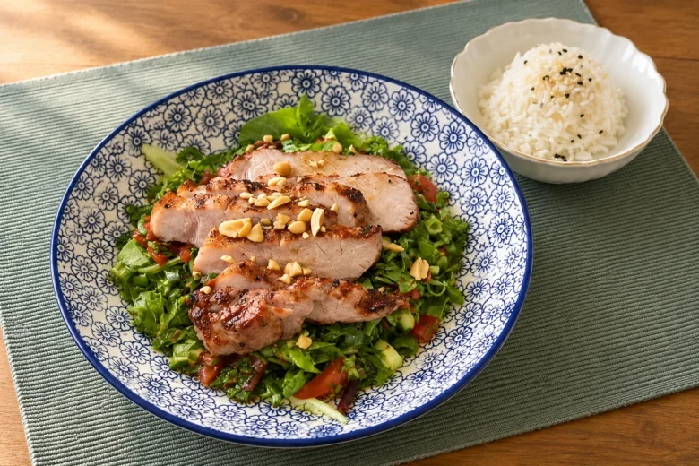 Sliced seared pork loin topped with chopped peanuts on a chopped green salad with tomato and cucumber, in a blue and white patterned bowl, with a small bowl of white rice sprinkled with sesame seeds beside it.
