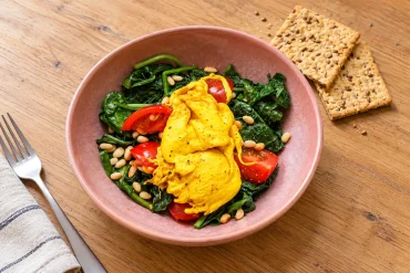 Turmeric poached eggs with spinach, cherry tomatoes and pine nuts in a pink bowl, with seeded crackers alongside.
