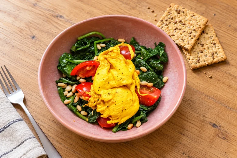 Turmeric poached eggs with spinach, cherry tomatoes and pine nuts in a pink bowl, with seeded crackers alongside.